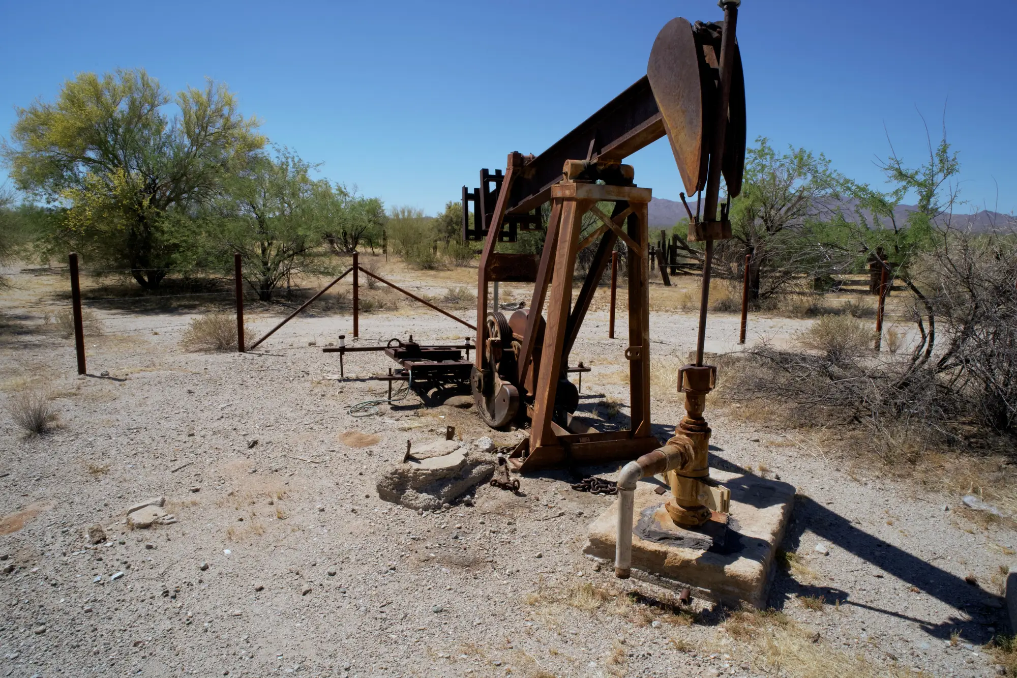 A rusted pumpjack on a dry, gravelled well pad with mesquite and scrub behind it